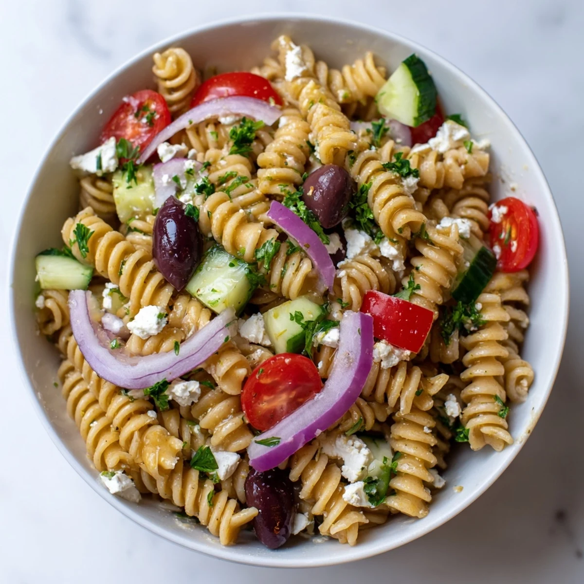 Fresh Mediterranean Pasta Salad with Olives in a white bowl, tossed with red cherry tomatoes, green cucumber slices, and creamy crumbled feta cheese.