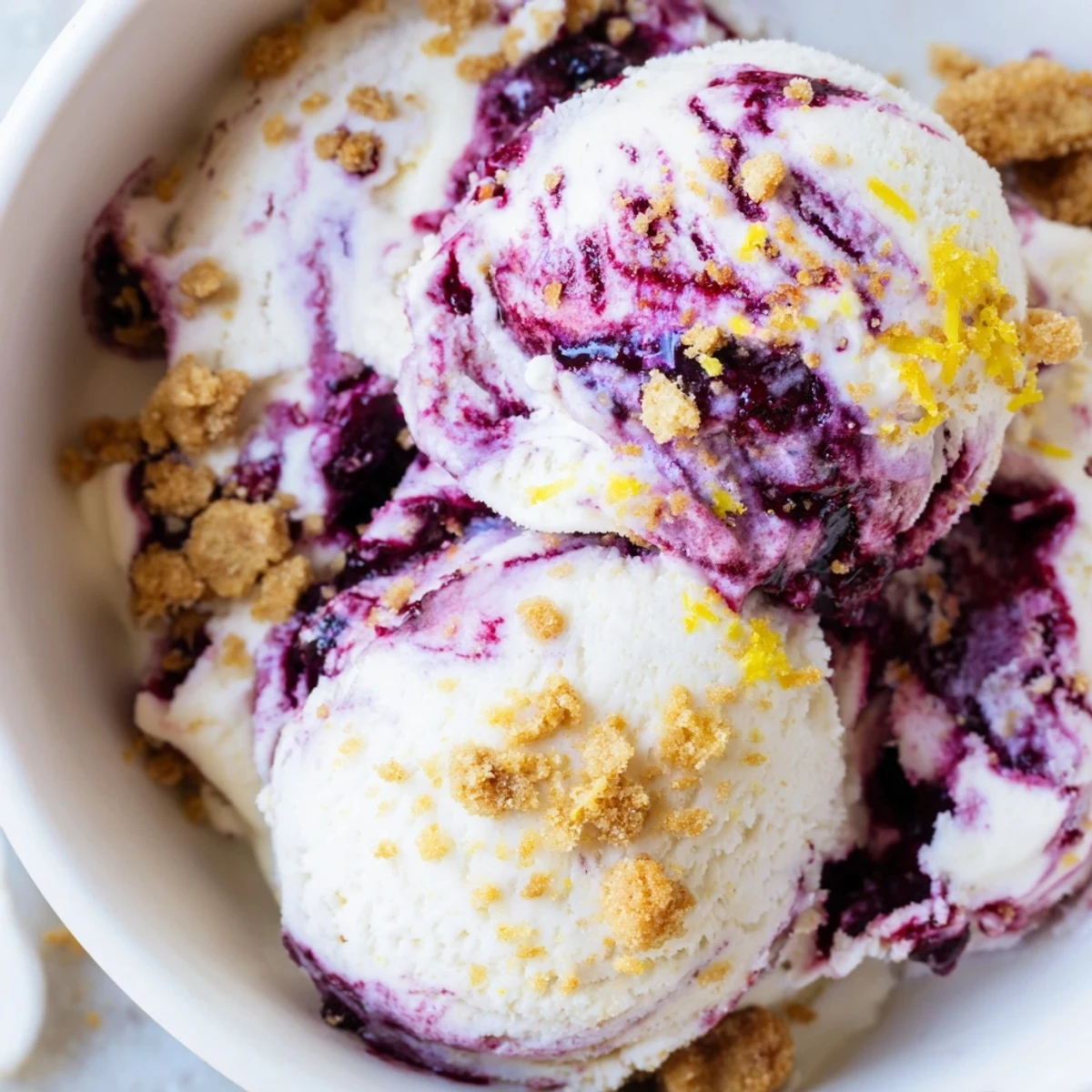 A scoops of Lemon Blueberry Cheesecake Ice Cream in a waffle cone with graham cracker crumbs.