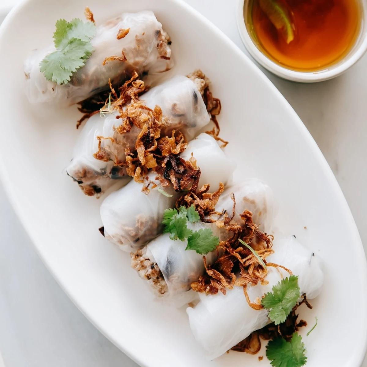 A platter of Banh Cuon topped with crispy shallots, cilantro, and bean sprouts alongside a bowl of nuoc cham.