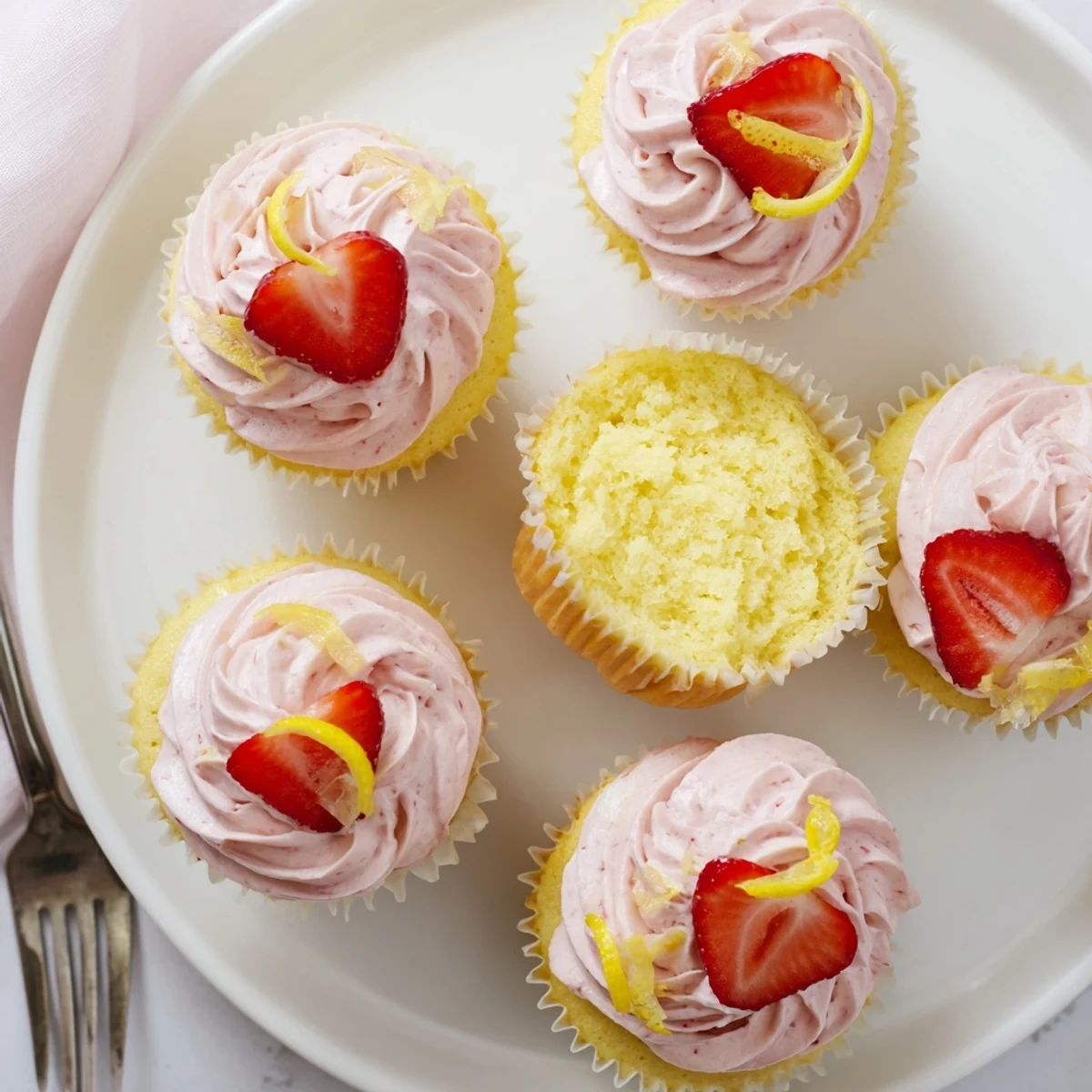 Golden lemon cupcakes topped with swirls of creamy pink strawberry buttercream frosting, garnished with fresh strawberry slices on a white cake stand.